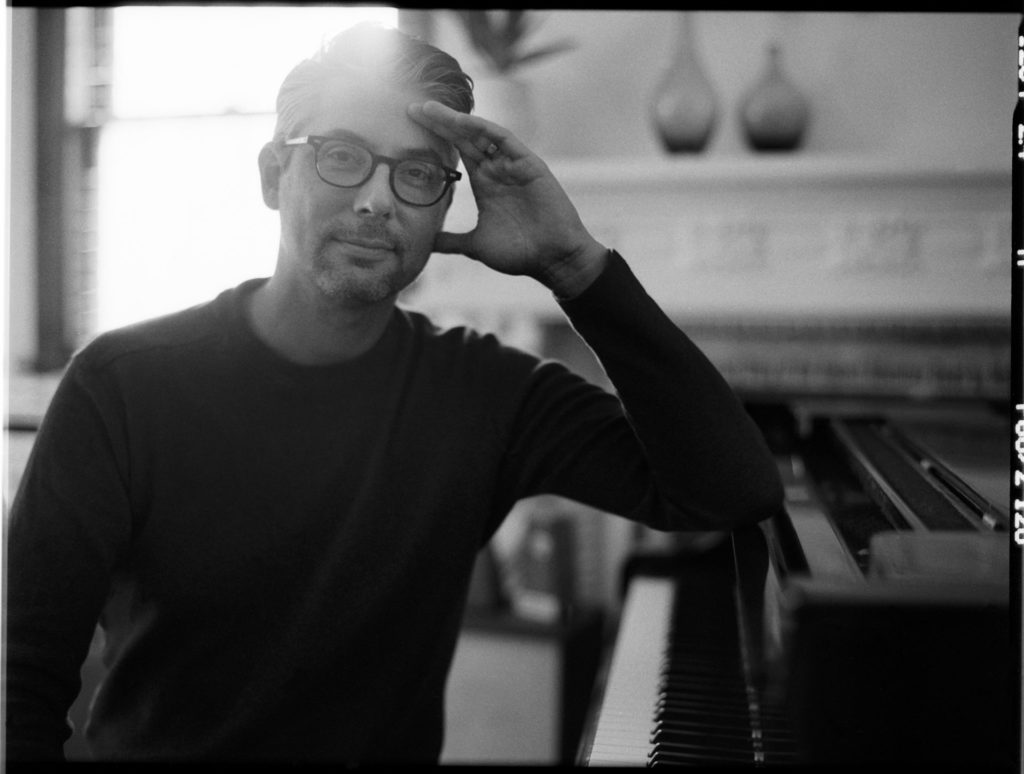 Josh Nelson in Black and White, leaning on a piano, posed and looking at the camera.
